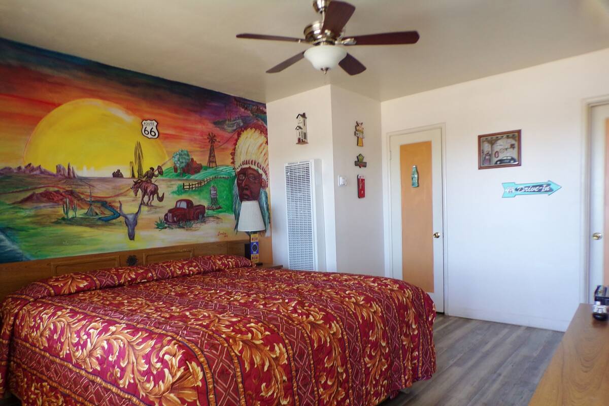 A bedroom with a large bed, a ceiling fan, a mural on the wall, and a door with a sign.