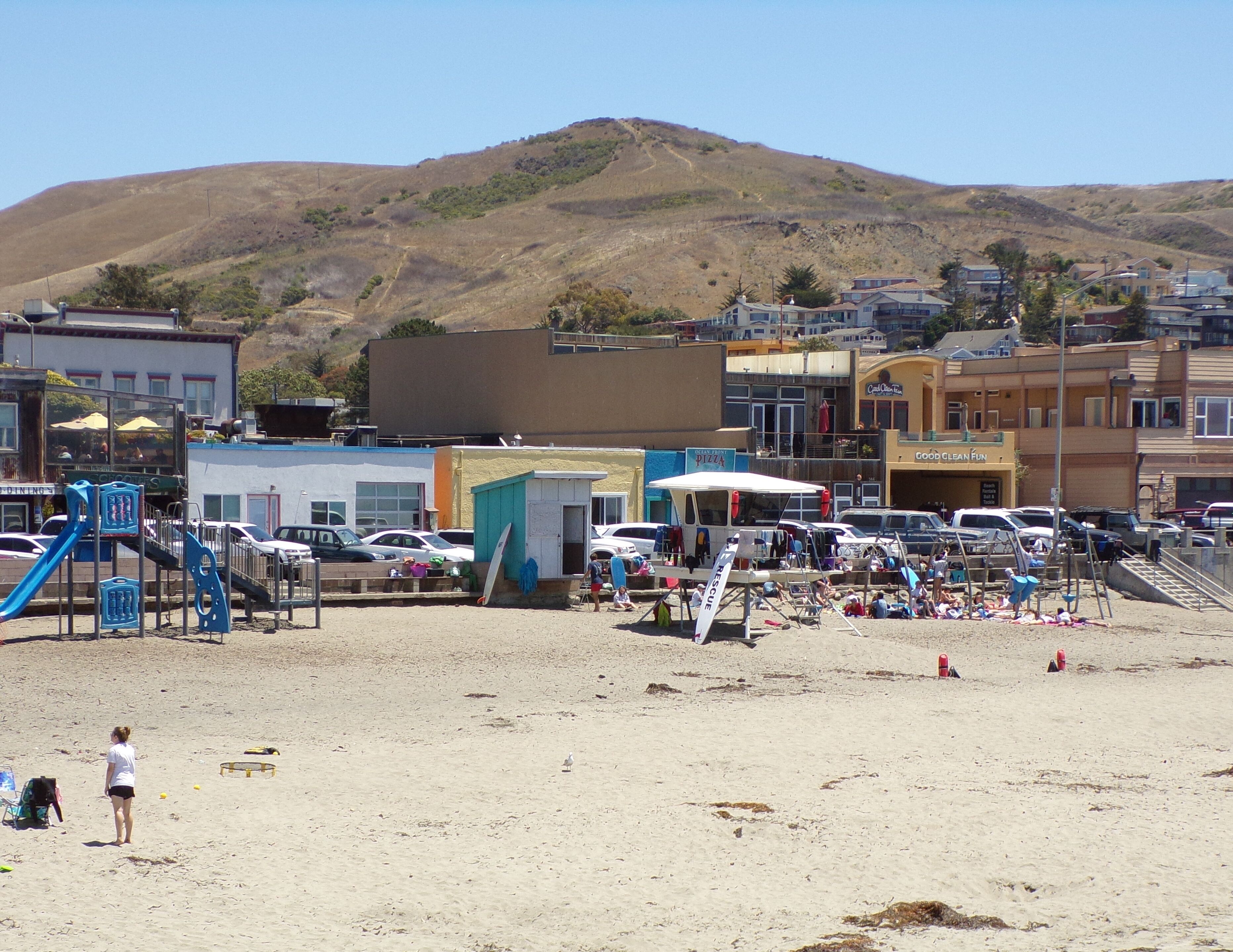 A beachside parking area with a playground, a small building, and a hill in the background.