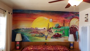 Een kamer met een bed, een plafondventilator, een muurschildering, een raam met gordijnen en een luchtverfrisser aan de muur.