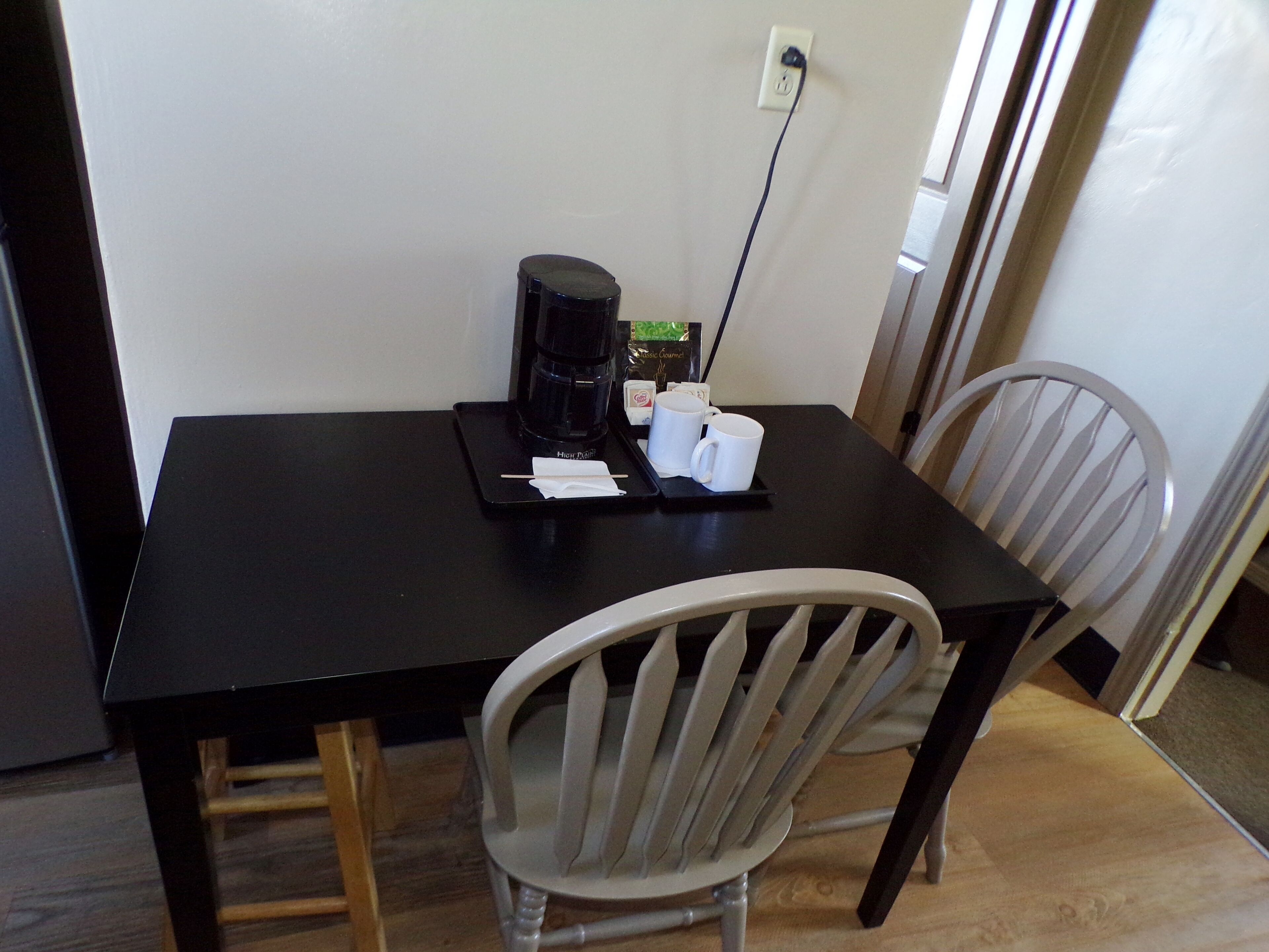 Une petite table de couleur sombre, sur laquelle se trouvent une machine à café, deux tasses blanches et un plateau avec une serviette. Une chaise blanche est placée devant la table.