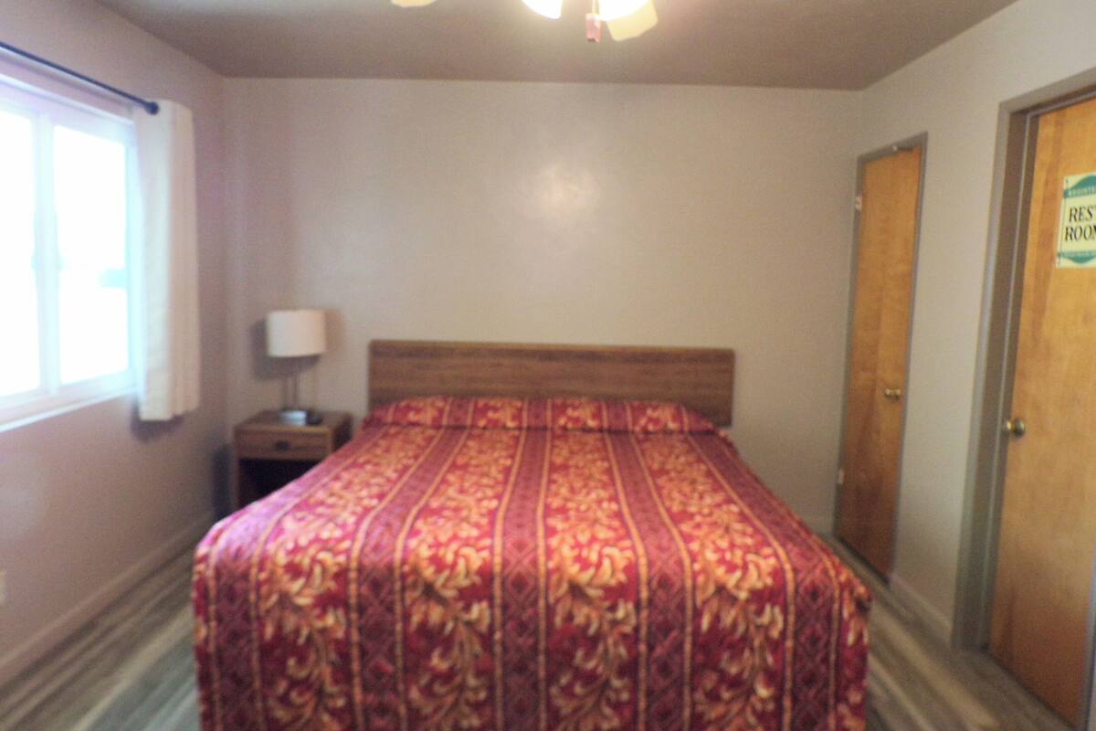 A bedroom with a bed, a nightstand, a ceiling fan, a window with curtains, and a door labeled "REST ROOM".