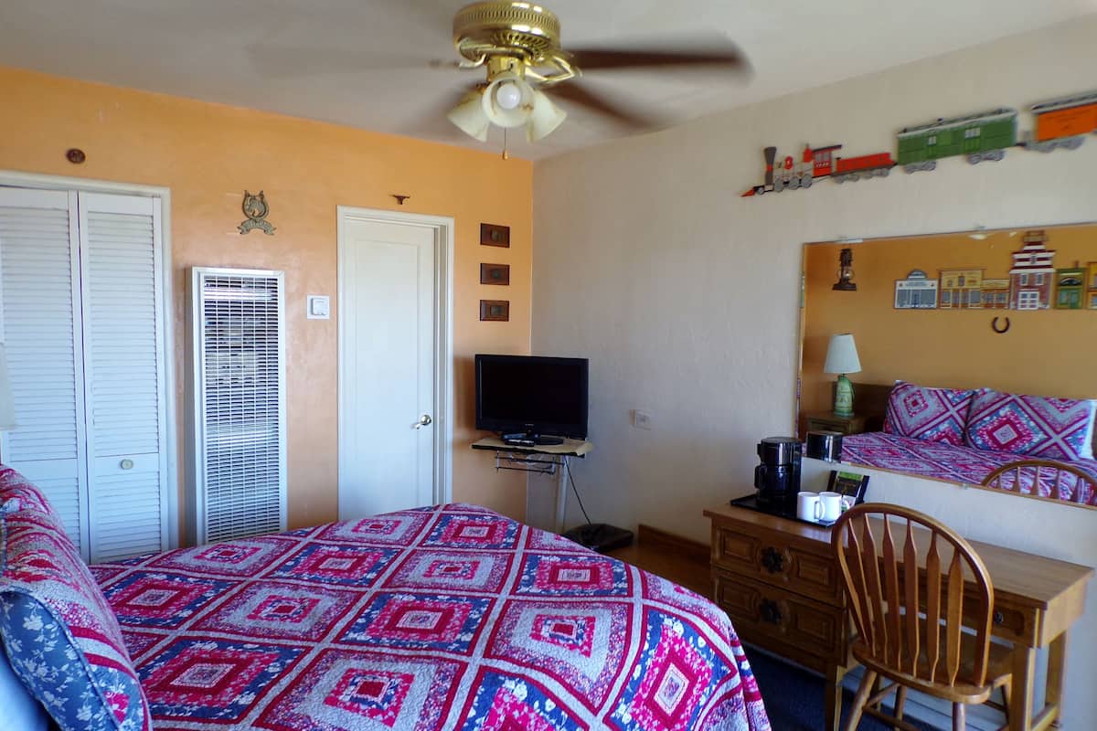 A bedroom with a bed, a desk, a chair, a television, a ceiling fan, and a wall with a mirror and decorations.