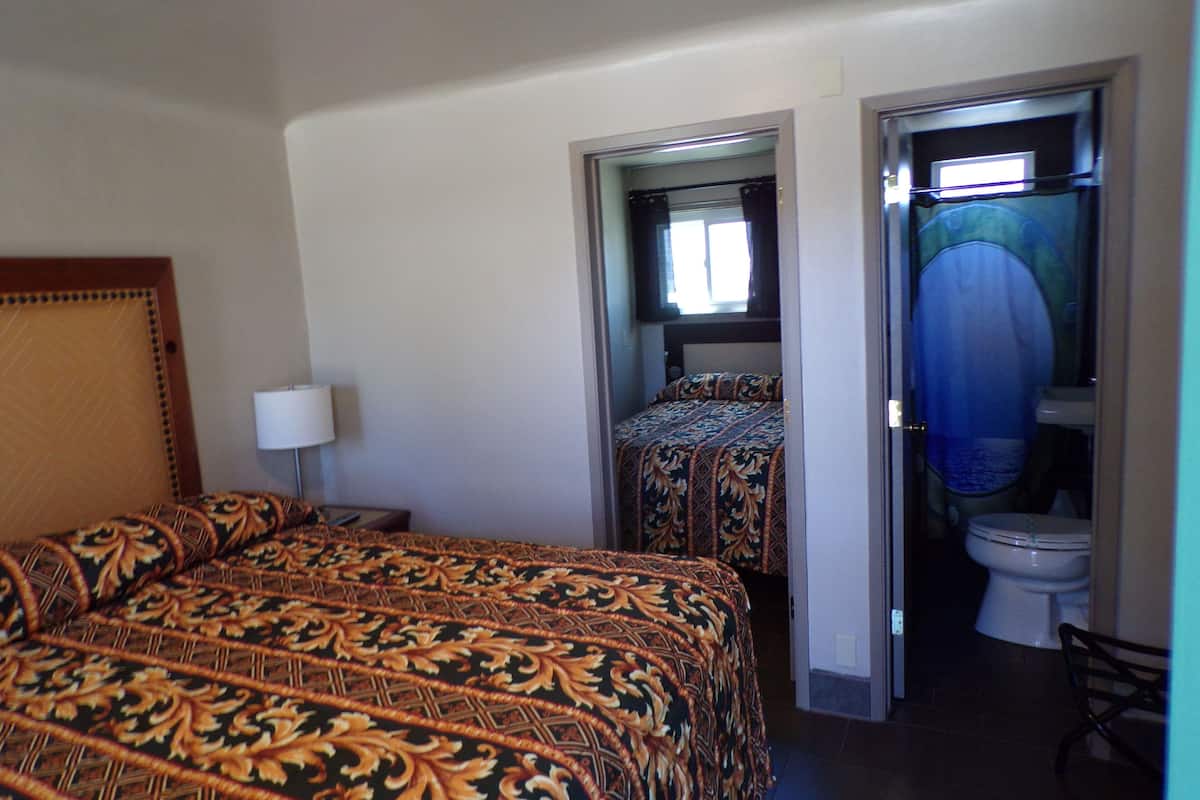 A hotel room with a large bed, a bedside table with a lamp, a bathroom with a toilet and shower, and a small window with curtains.