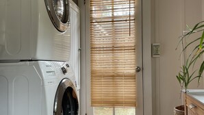 A laundry room with stacked washers and dryers, a door with blinds, a wooden floor, and a wicker basket. - Cozy get-away in Pacific Grove (Pet Friendly!) (Pacific Grove)