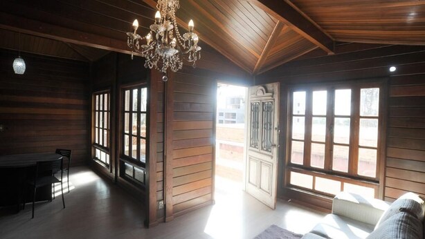 Um quarto amplo com paredes de madeira, um lustre, janelas grandes e uma porta que dá para o exterior.