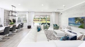 A modern living room with a white sofa, a dining area with a long table and chairs, a TV on the wall, and a view of the outdoors. - 8 Bdrm Villa w/Pool, Theater Near Disney w/ Waterpark & Free Shuttle Service (Kissimmee)