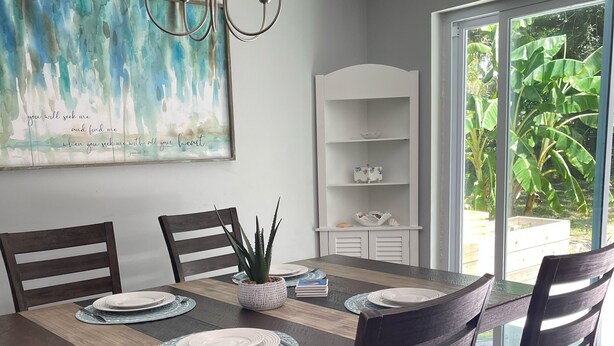 Un coin repas avec une table en bois dressée pour quatre personnes, entourée de chaises en bois foncé. Un lustre est suspendu au-dessus, et un grand tableau abstrait orne le mur.