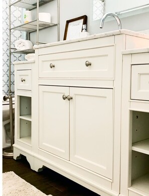 A white vanity with drawers and a cabinet, a shelf with toiletries, and a framed quote above the mirror.