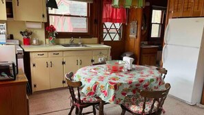 A kitchen with a round table covered in a floral tablecloth, wooden chairs, a refrigerator, and a window with a view of a house. - Cheerful cottage with 4 private beach accesses!!!! (East Tawas)