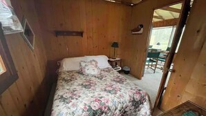 A wooden cabin room with a bed, a small table, a lamp, and a view of the outside. - Cheerful cottage with 4 private beach accesses!!!! (East Tawas)
