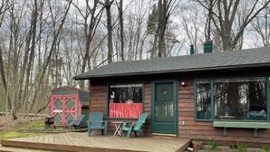 A wooden cabin with a porch, two chairs, and a red barn-like structure in the background. - Cheerful cottage with 4 private beach accesses!!!! (East Tawas)