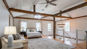 A spacious bedroom with a large bed, a sitting area with a sofa and a lamp, wooden beams on the ceiling, and a staircase with a railing.