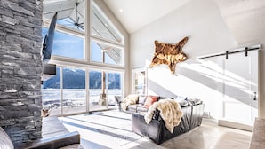 A spacious living room with a stone wall, large windows, a leather sofa, and a mounted animal hide. - New Lakefront Modern Chalet With Panoramic Views (Donnelly)