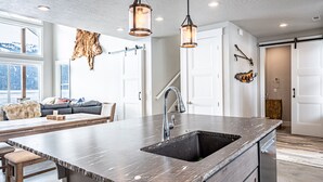 A modern kitchen with a large island, stainless steel sink, and pendant lighting. - New Lakefront Modern Chalet With Panoramic Views (Donnelly)