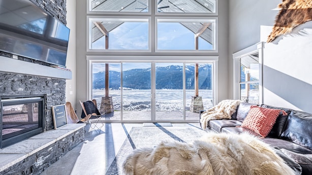 A spacious living room with a stone fireplace, a large window offering a view of a snowy landscape, and a leather sofa with a fur throw. - New Lakefront Modern Chalet With Panoramic Views (Donnelly)