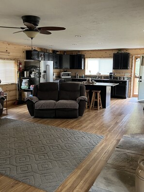 L’intérieur d’un chalet accueillant présente un salon avec un canapé de couleur foncée, un tapis à motifs et un plancher en bois. On aperçoit à l’arrière-plan une cuisine dotée de meubles noirs et d’un îlot central.