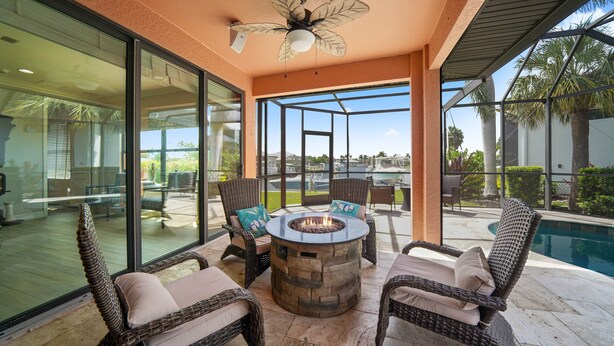 A patio area with wicker furniture, a stone fire pit, and a glass enclosure overlooking a pool and greenery. - Beautiful Gulf Access home Amazing pool sunsets  (Punta Gorda)