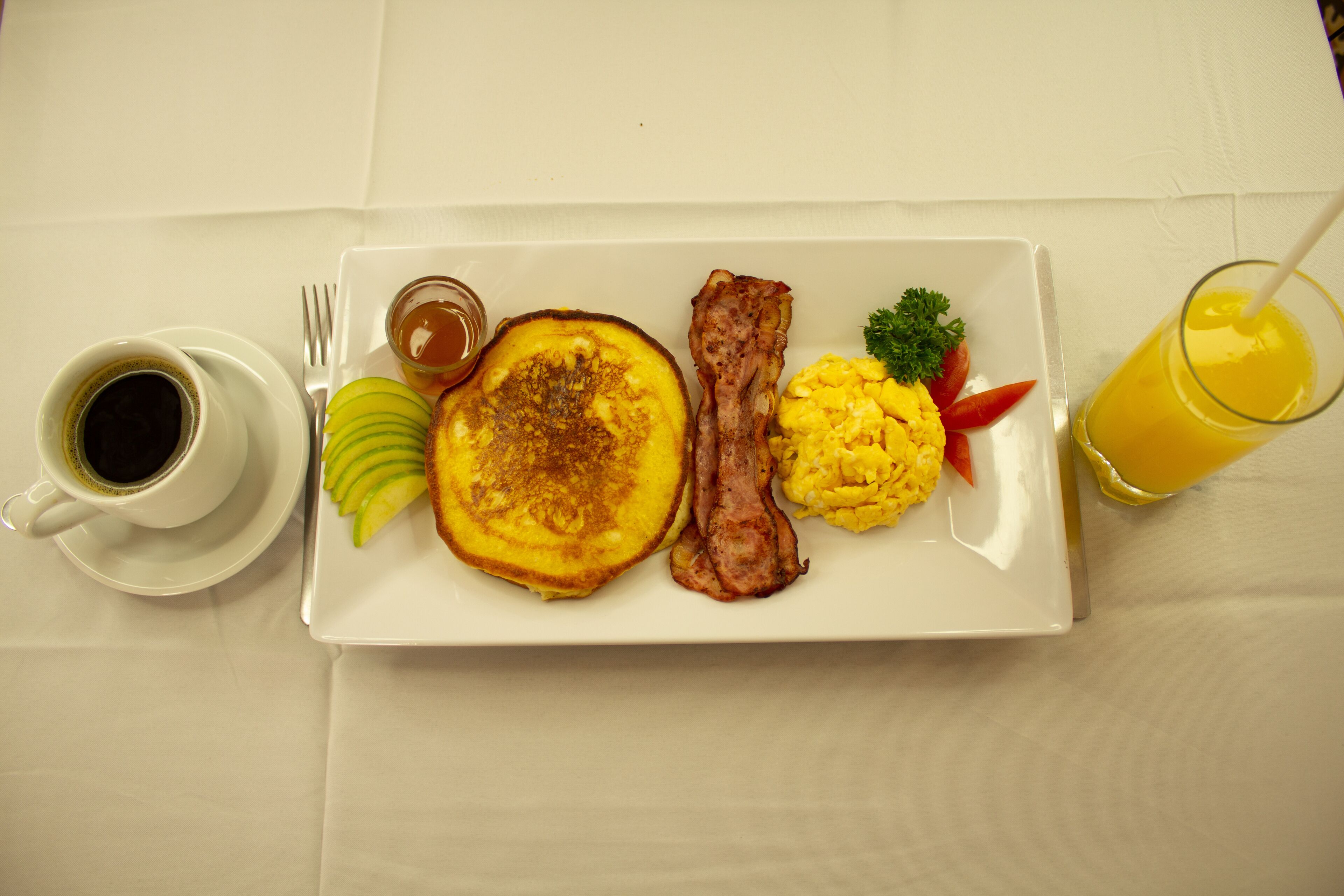 En morgenmadstallerken med pandekager, bacon, røræg og et glas appelsinjuice.