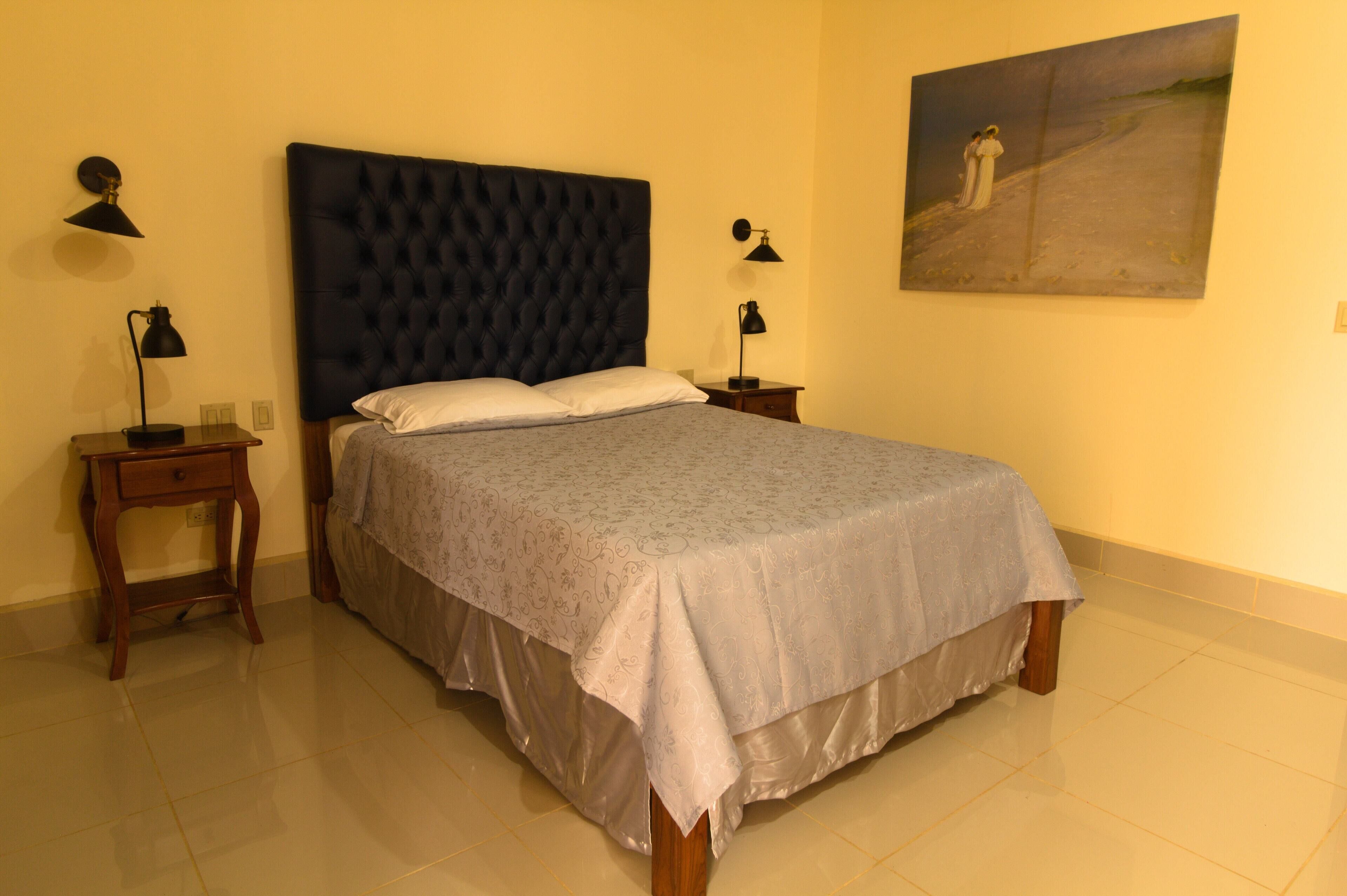 A bedroom with a tufted headboard, a bed with a patterned bedspread, two bedside tables with lamps, and a painting on the wall.