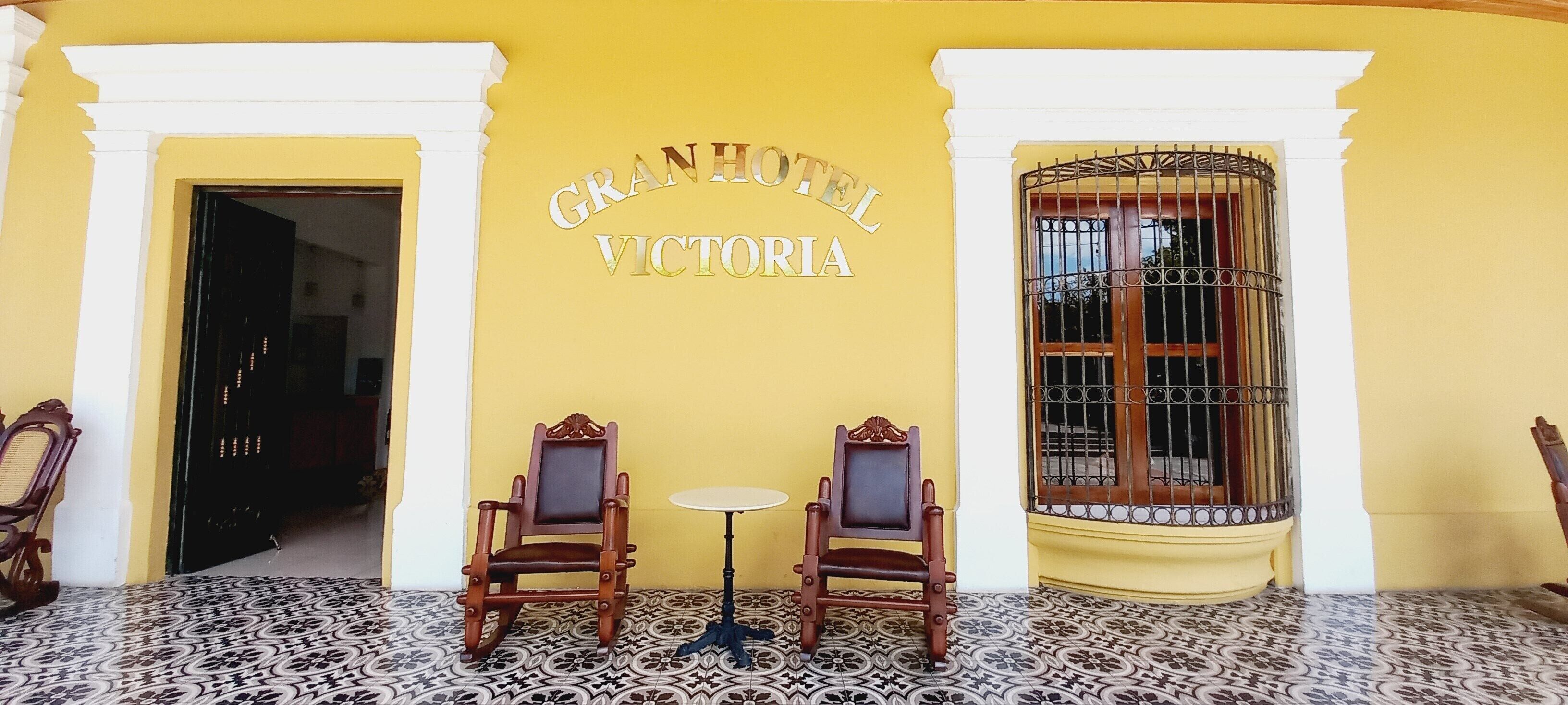 Exterior del Gran Hotel Victoria con dos sillas de madera y una pequeña mesa del frente, piso de baldosas y un amplio ventanal con reja de metal.