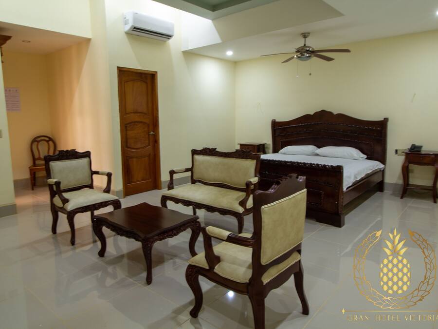 A hotel room with a wooden bed, two upholstered chairs, a small table, and a ceiling fan.