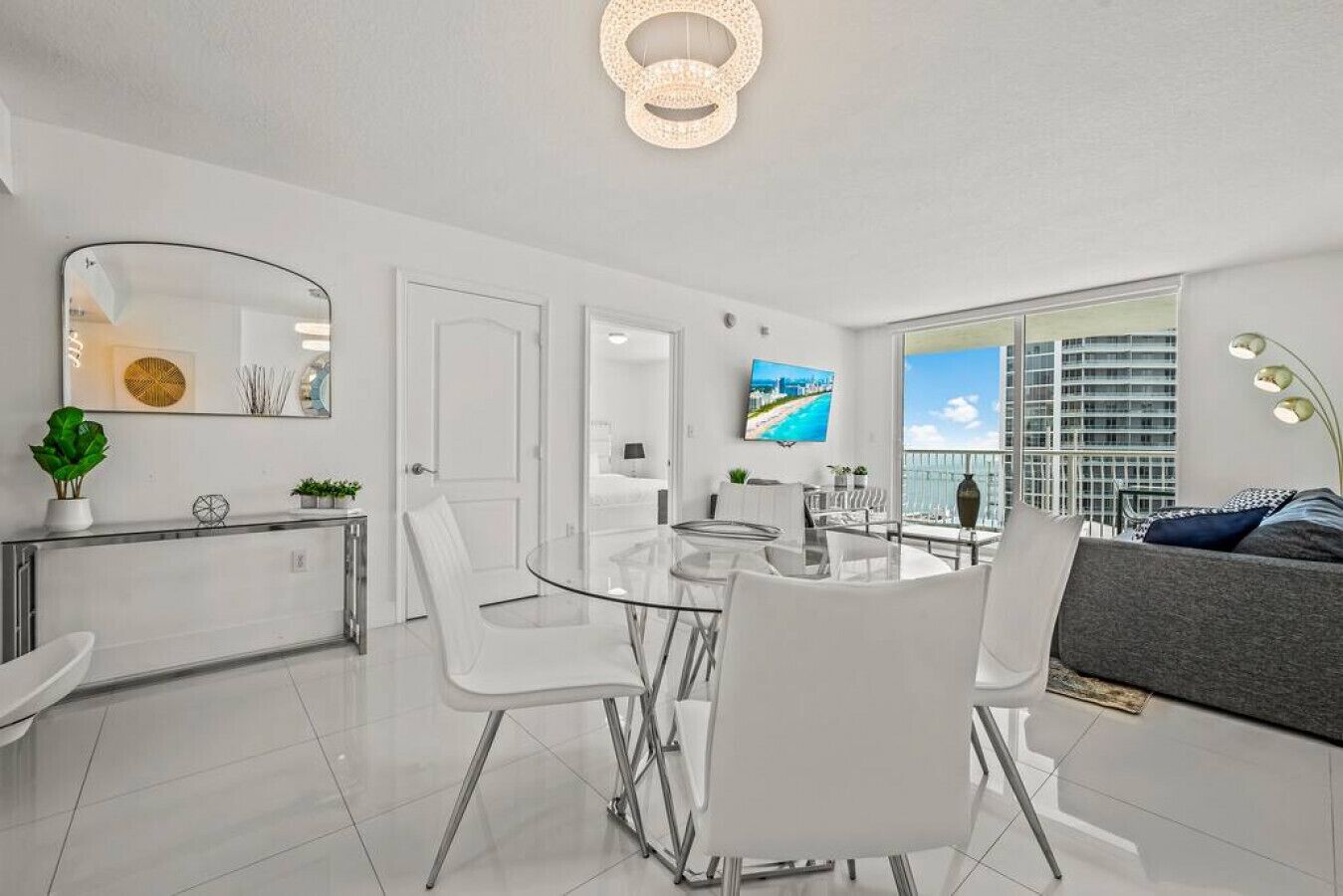 A modern, well-lit living room with a glass dining table, white chairs, a grey sofa, and a large window offering a city view.