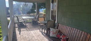 Eine überdachte Terrasse mit Tisch und Stühlen, eine Holzbank mit Kissen und Blick auf eine Stadtlandschaft durch große Fenster.