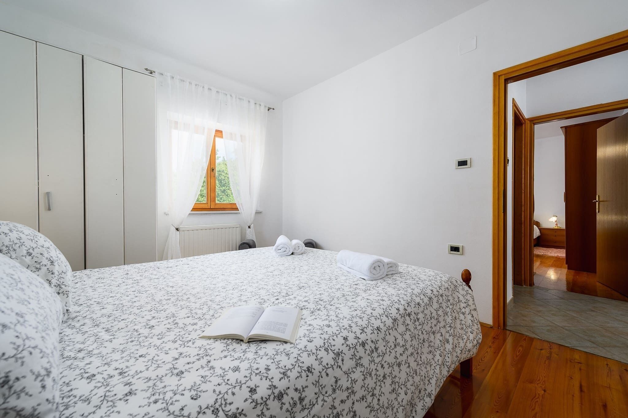 A bedroom with a bed, a window with curtains, a radiator, and a wooden door leading to another room.