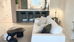 Een moderne woonkamer met een witte bank, een bijzettafeltje en een gevlochten mand. In de achtergrond is een keuken met een bar en hanglampen zichtbaar.