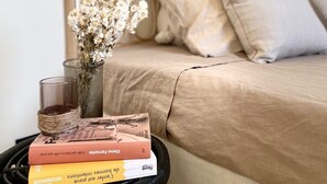 Een bed met een beige deken, een vaas met gedroogde bloemen, een glazen kandelaar en een stapel boeken op een ronde zwarte tafel.