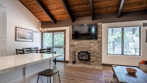A modern kitchen with a stone fireplace, wooden ceiling beams, and a large TV mounted above it.