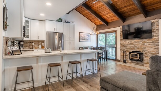 A modern kitchen with white cabinets, a large island with stools, a built-in stove, and a stone fireplace.