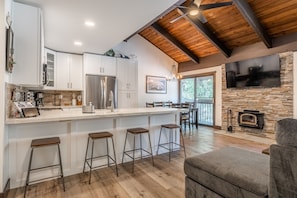 A modern kitchen with white cabinets, a large island with stools, a built-in stove, and a stone fireplace.