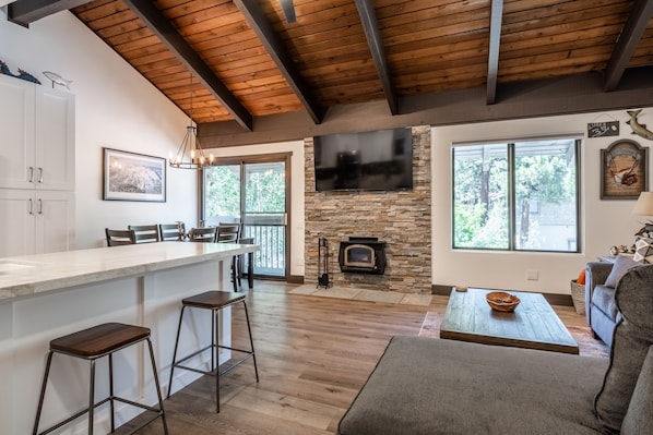A spacious living area with a stone fireplace, a large TV mounted on the wall, and a wooden ceiling.