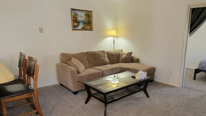 Uma sala de estar com um sofá bege, uma mesa de centro de vidro, uma cadeira de madeira e um abajur.