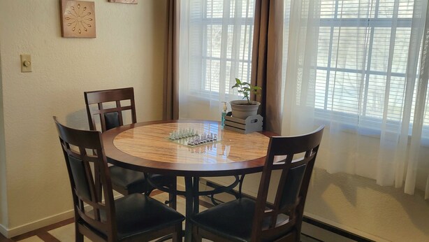 Uma área de refeições com uma mesa redonda, quatro cadeiras, uma luminária pendente e uma janela com cortinas.