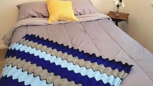 Um quarto com cama, travesseiro, cobertor, mesa de cabeceira com um vaso e um quadro na parede.
