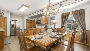 Comedor con una mesa de madera dispuesta para seis comensales, rodeada de sillas de madera. Un candelabro cuelga sobre la mesa, y se ve una ventana con cortinas a la derecha.