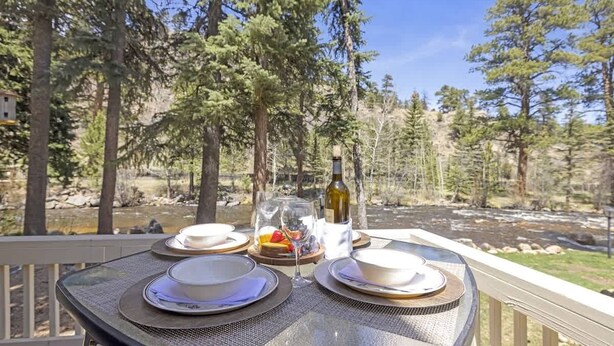 Mesa preparada para dos personas, con platos, vasos y una botella de vino, con vista a un río y una zona boscosa.