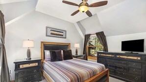 Un dormitorio con una cama grande, dos mesitas de noche, un aparador, un ventilador de techo, un televisor y una ventana con cortinas.