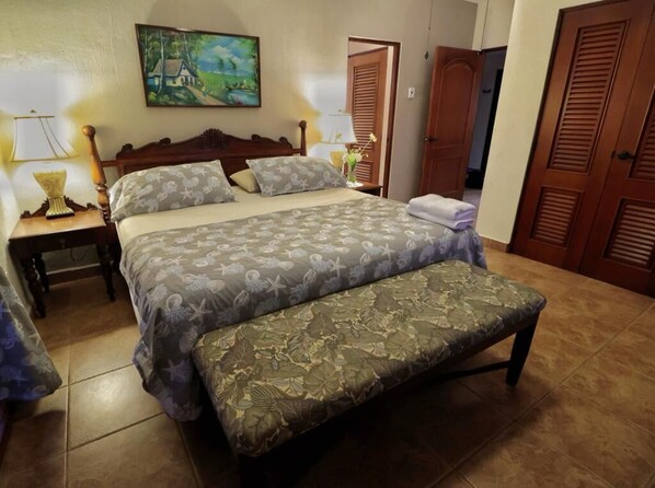 Un dormitorio con una cama, un banco, una mesita de noche, una lámpara, un cuadro y puertas de madera.