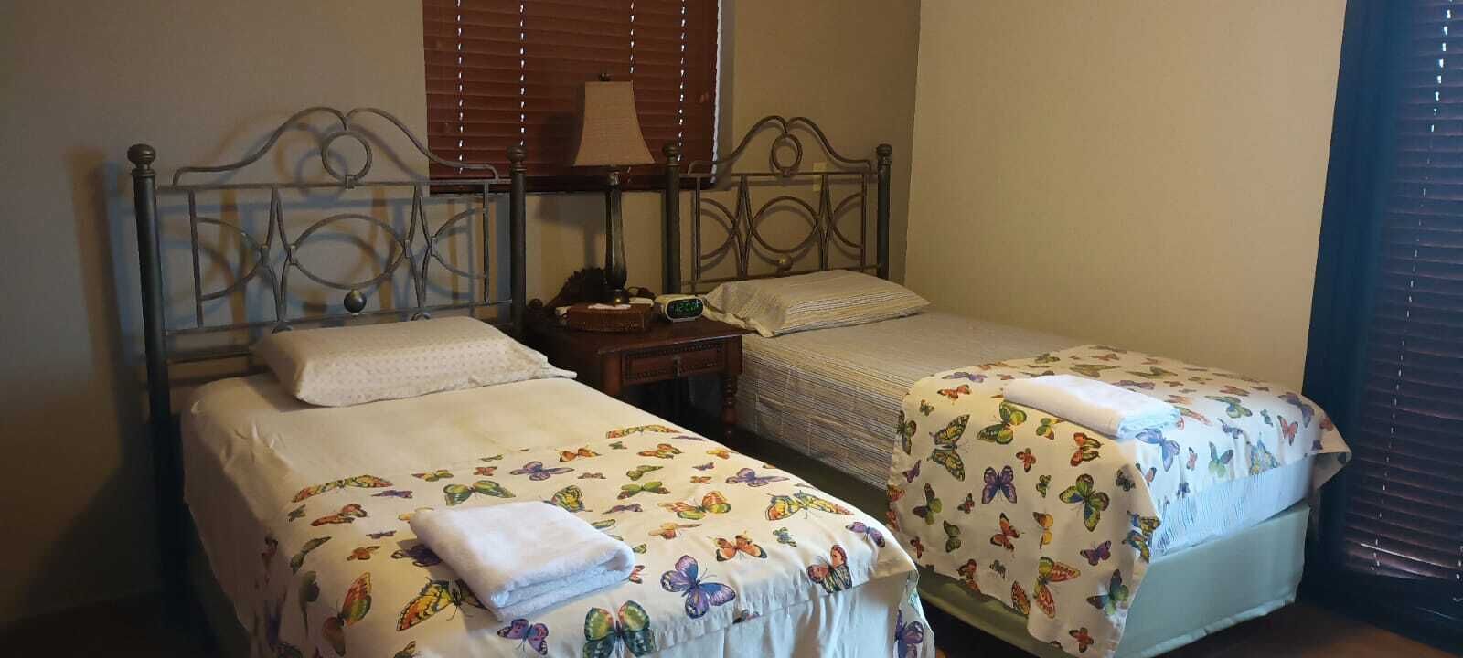 Une chambre avec deux lits simples, chacun recouvert d’une couvre-lit à motifs floraux et agrémenté d’une serviette blanche pliée. Une table de chevet avec une lampe et un réveil se trouve entre les deux lits.