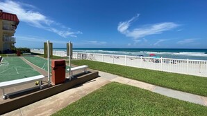 A sunny day at a beachside location with a shuffleboard court, a white fence, and a view of the ocean.