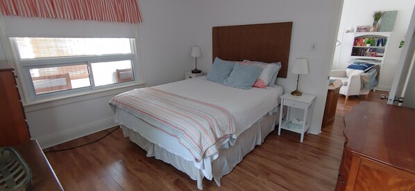 A bedroom with a large bed, two bedside tables, a nightstand, a wooden dresser, a window with blinds, and a bookshelf with books and decorative items.