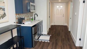 A compact kitchen with dark blue cabinets, a white countertop, and a microwave. A small black table is positioned near the counter. The floor is wooden, and a white door is visible in the background. - Historic St. Augustine Balcony Apartment (Saint Augustine)