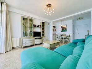 A living room with a teal sofa, a TV on a cabinet, a dining table with chairs, and a chandelier.