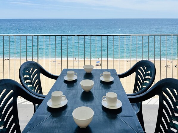 A table with white cups and bowls, black chairs, and a view of the beach and ocean.
