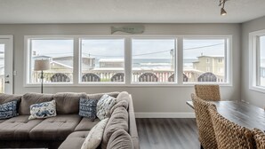 A living room with a sofa, a dining table, and three large windows offering a view of the beach and neighboring houses.