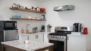 A compact kitchen with a microwave, stove, oven, and coffee maker. There are wooden shelves above the counter holding kitchenware. - Small Studio - Trinity Groves - 5 Min Downtown (Dallas)