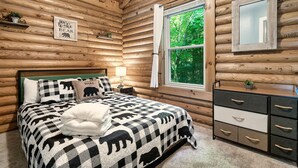Ein Schlafzimmer in einer Blockhütte mit einem Bett, das mit einer schwarz-weiß gestreiften Bären-Steppdecke ausgestattet ist, einem Nachttisch mit Lampe, einem Fenster mit Blick auf Grünflächen und einer Kommode mit Schubladen.